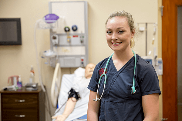 Female nursing student looking at the camera wearing gray scrubs standing in front of simulation mannequin